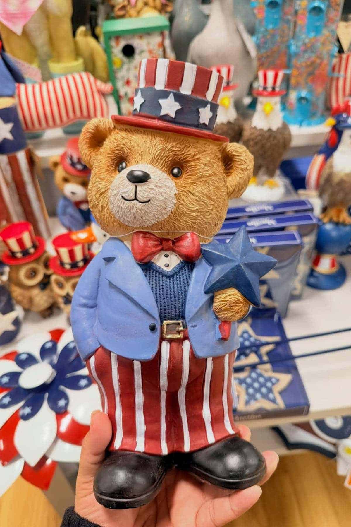 A hand holds a bear figurine dressed in a patriotic Uncle Sam outfit, with a blue jacket, red and white striped pants, and holding a blue star. Other similar decorations are in the background.