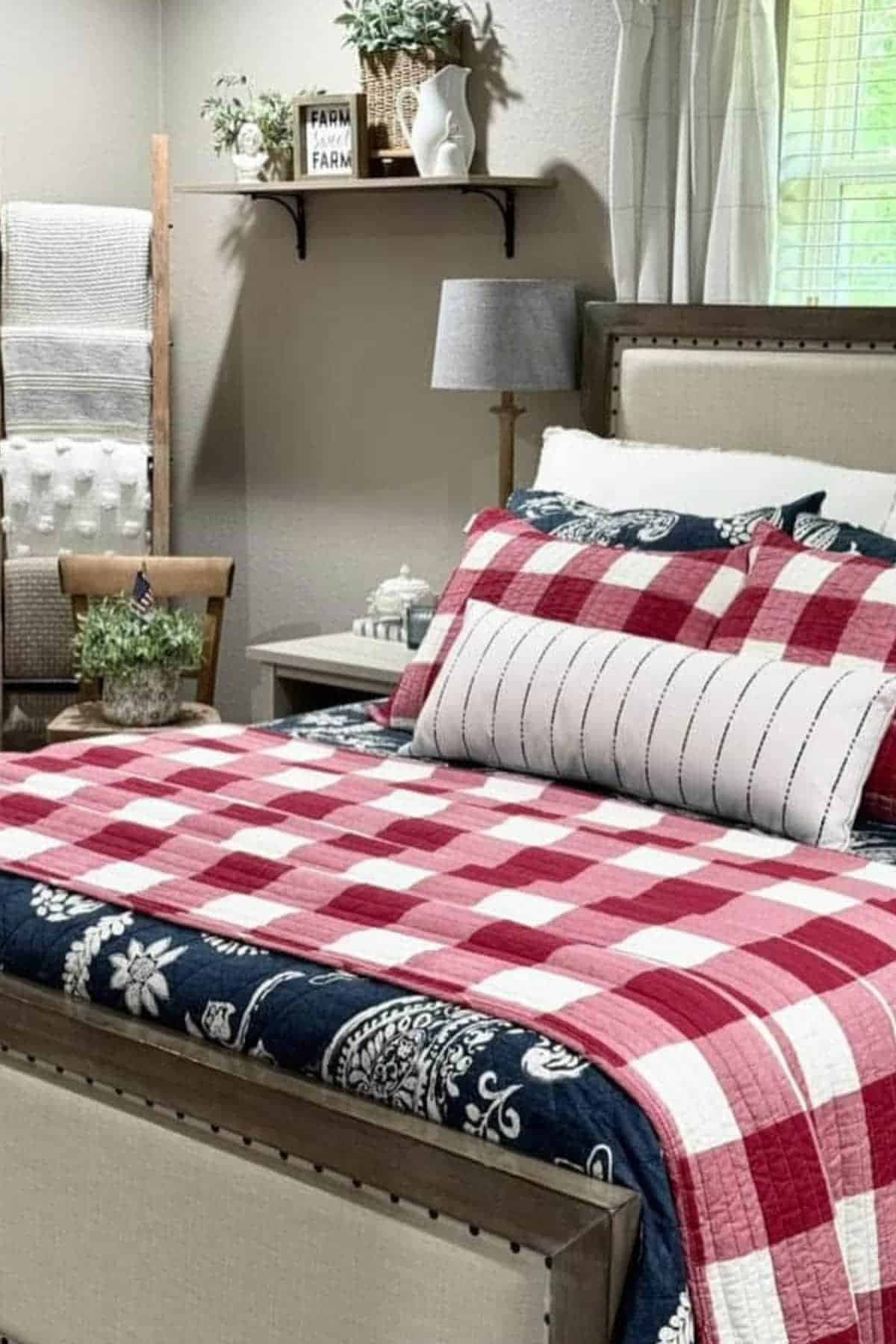 A neatly made bed with a red and white patchwork quilt, decorative pillows, and a small plant on a bedside table in a cozy, well-lit bedroom.