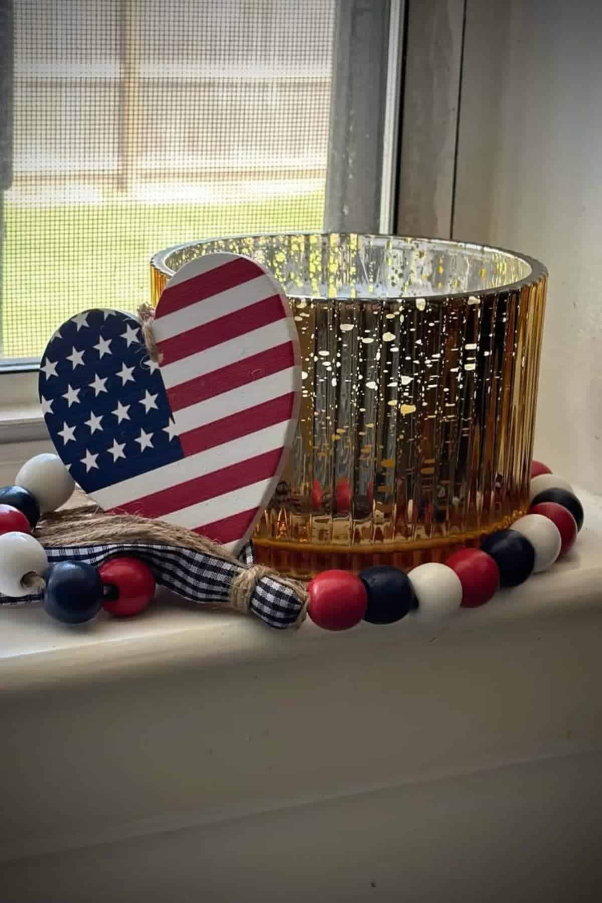 A gold glass candle holder sits on a windowsill, next to a wooden heart with an American flag design and a red, white, and blue bead garland.
