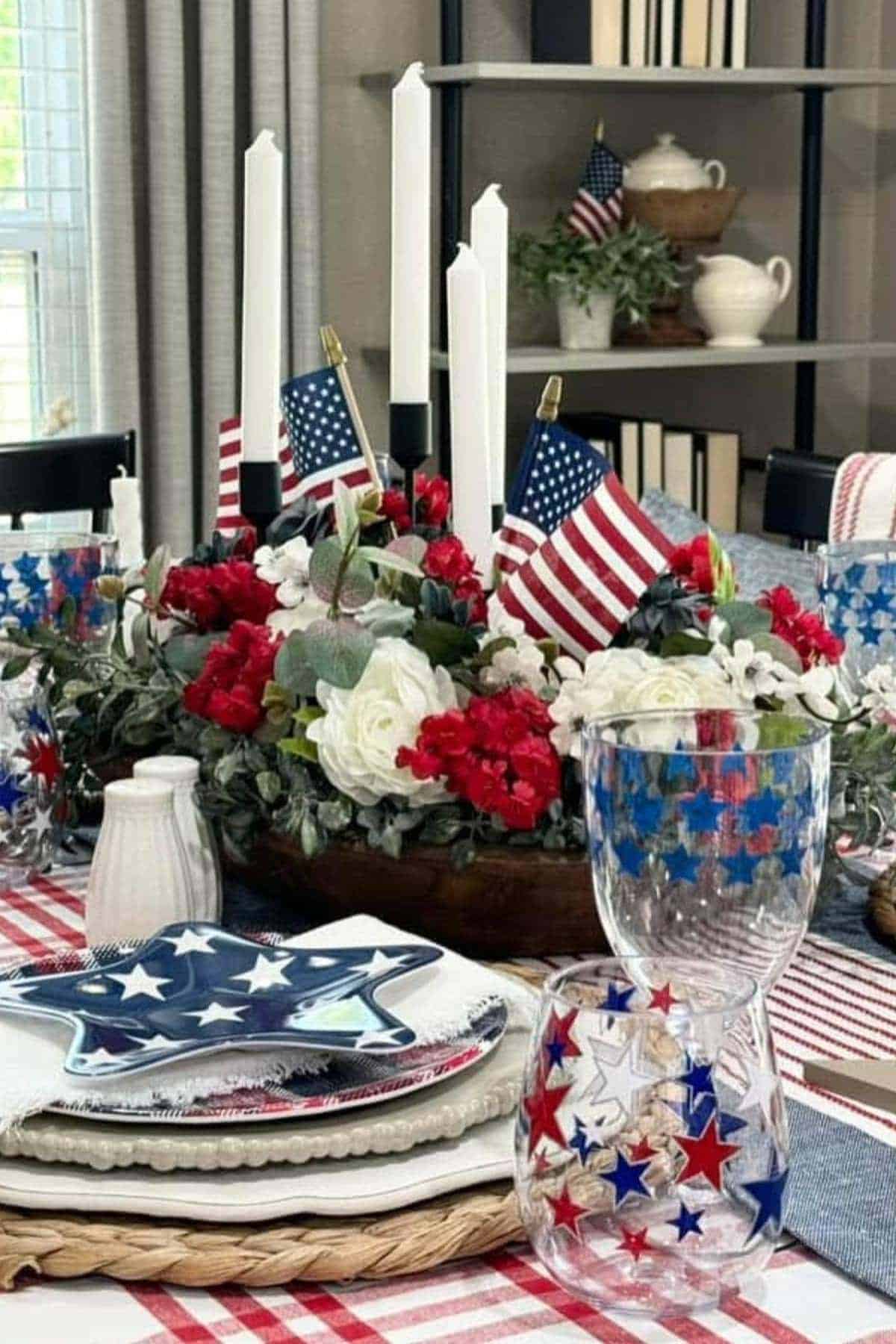 A dining table set with American flag decorations, red, white, and blue flowers, star-patterned glasses, and white candles on a checkered tablecloth.