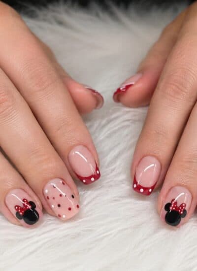 Hands with red and white polka dot and Minnie Mouse-themed nail art, resting on a white textured surface. Two gold rings are worn on each hand.