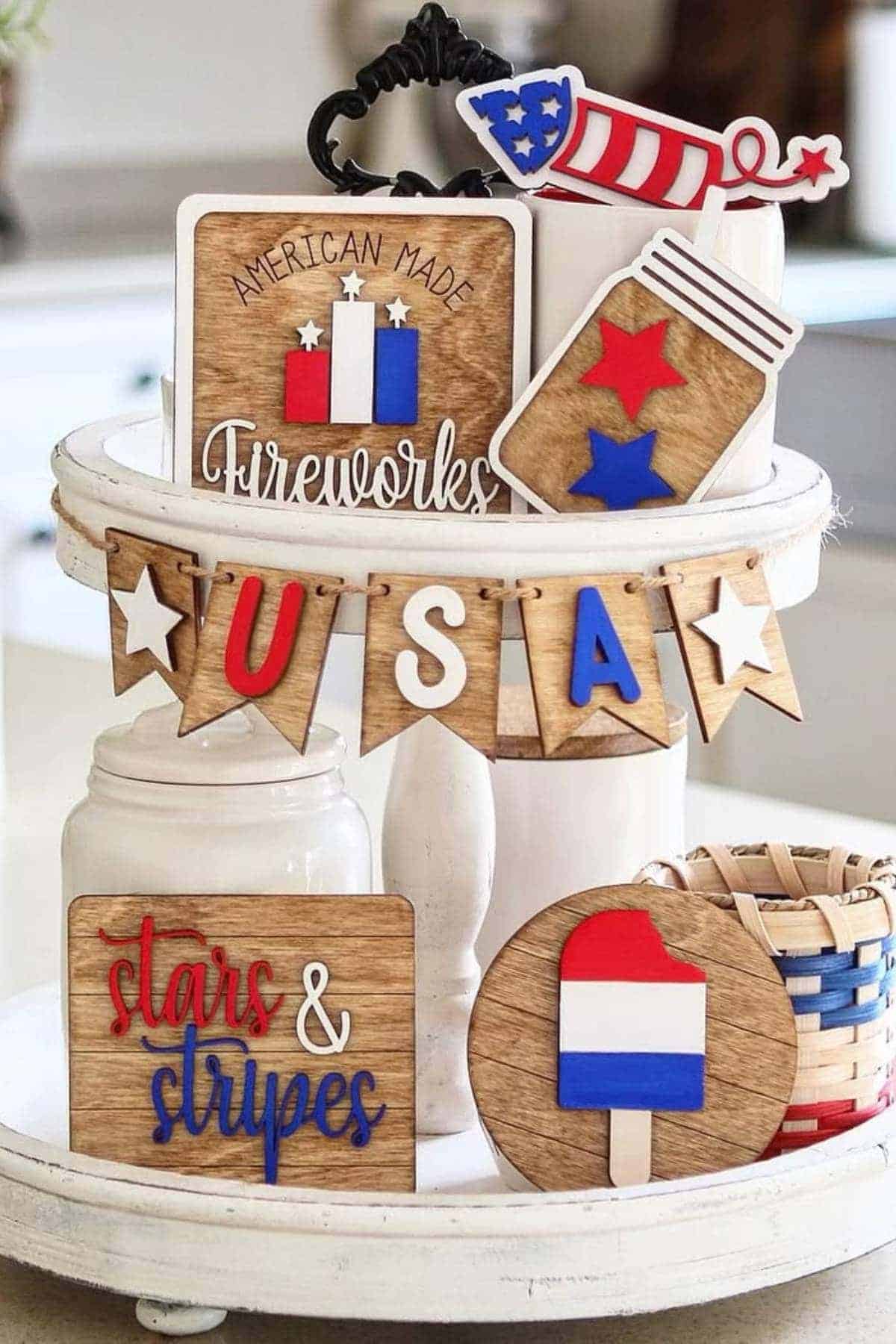 A two-tiered tray holds wooden decorations with patriotic themes, including "USA," stars, a popsicle, fireworks, and "stars & stripes," in red, white, and blue colors.