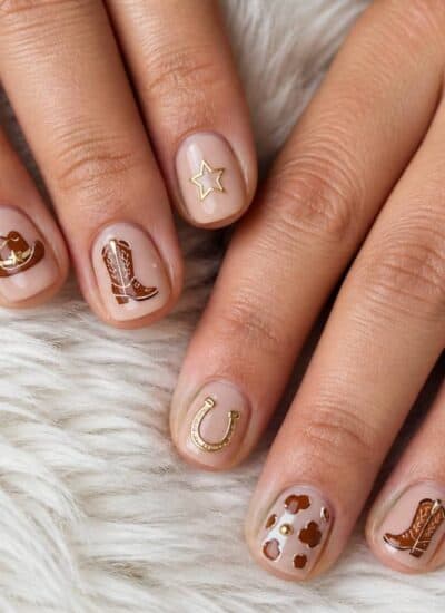Hands with short, nude-colored nails featuring western-themed nail art designs such as boots, a hat, a horseshoe, stars, and a key, placed on a white furry surface.