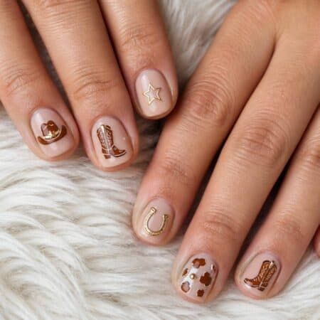 Hands with short, nude-colored nails featuring western-themed nail art designs such as boots, a hat, a horseshoe, stars, and a key, placed on a white furry surface.