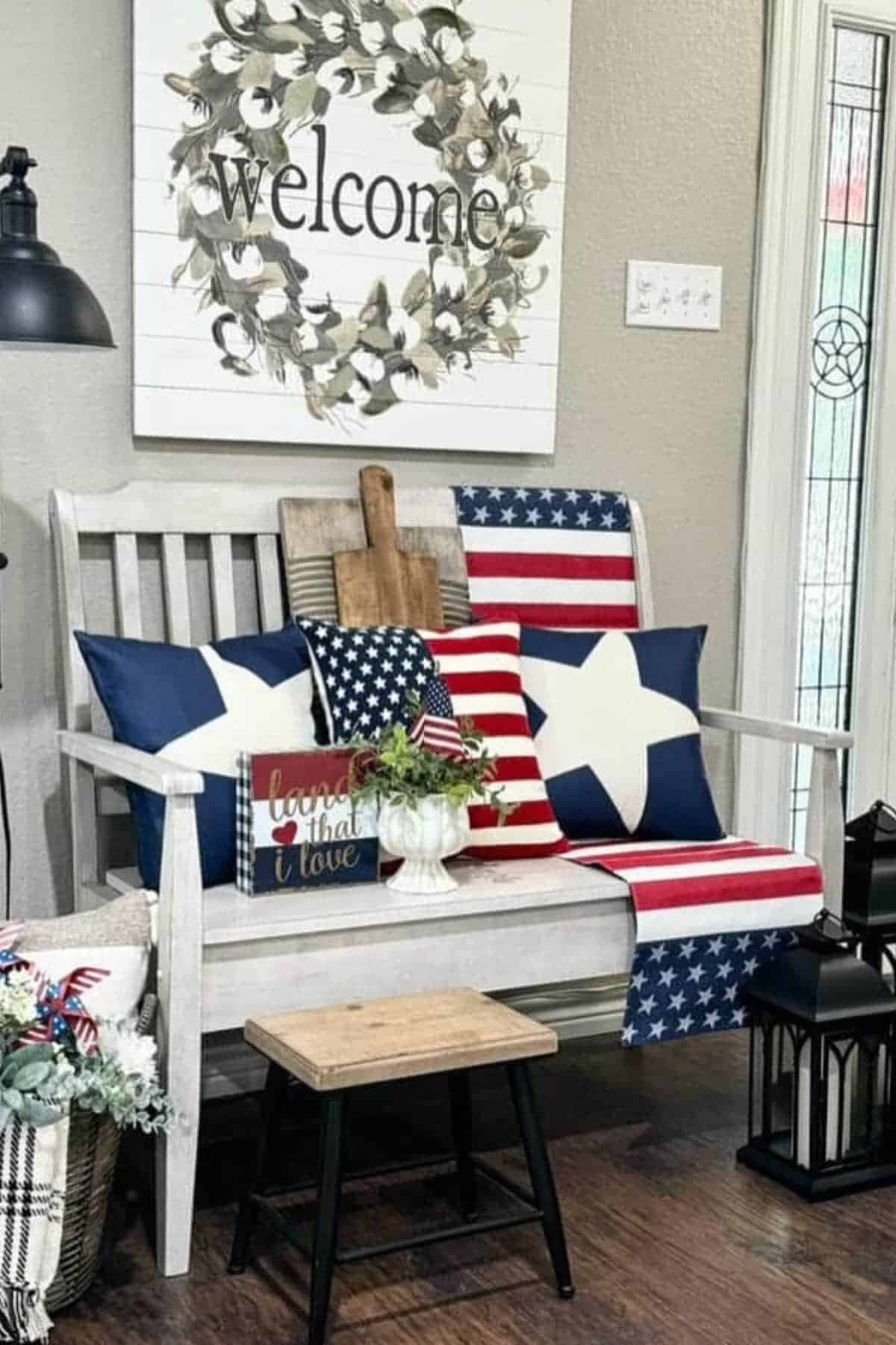 A white wooden bench with patriotic star and striped pillows, a small American flag, and a “welcome” sign above, set in a cozy, rustic home entryway.