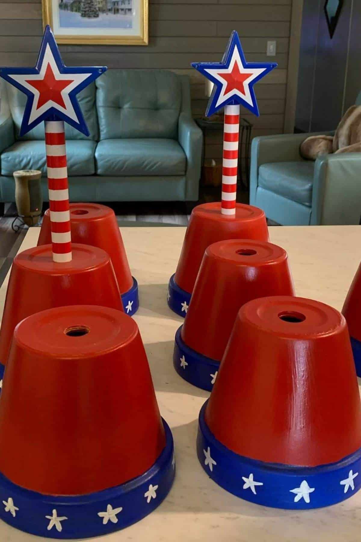 Four red flower pots painted with blue and white stars, topped with striped poles and star shapes, are arranged on a table in a living room.