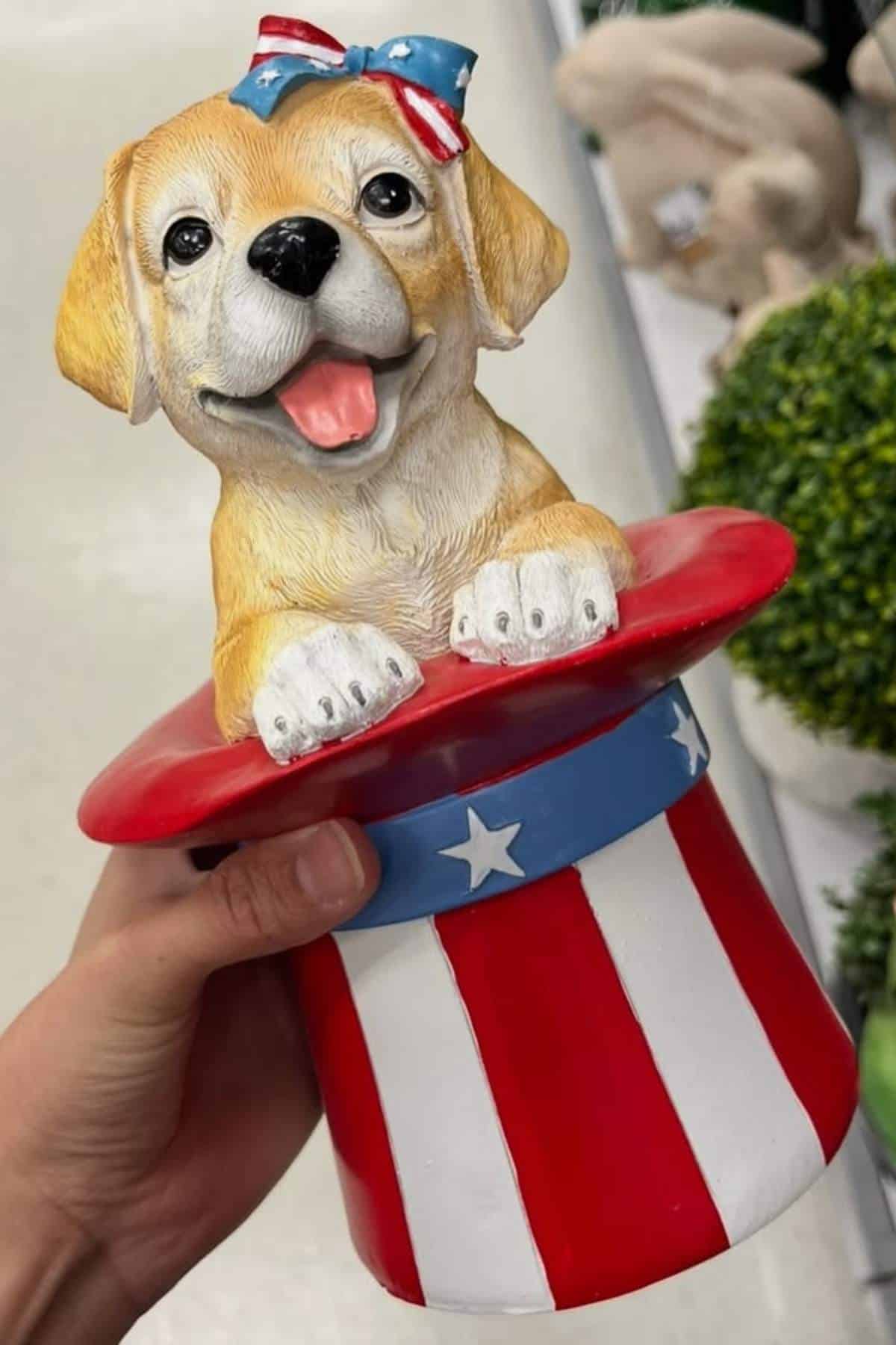 A hand holds a decorative figurine of a puppy wearing a bow, emerging from a patriotic hat with stars and red, white, and blue stripes.