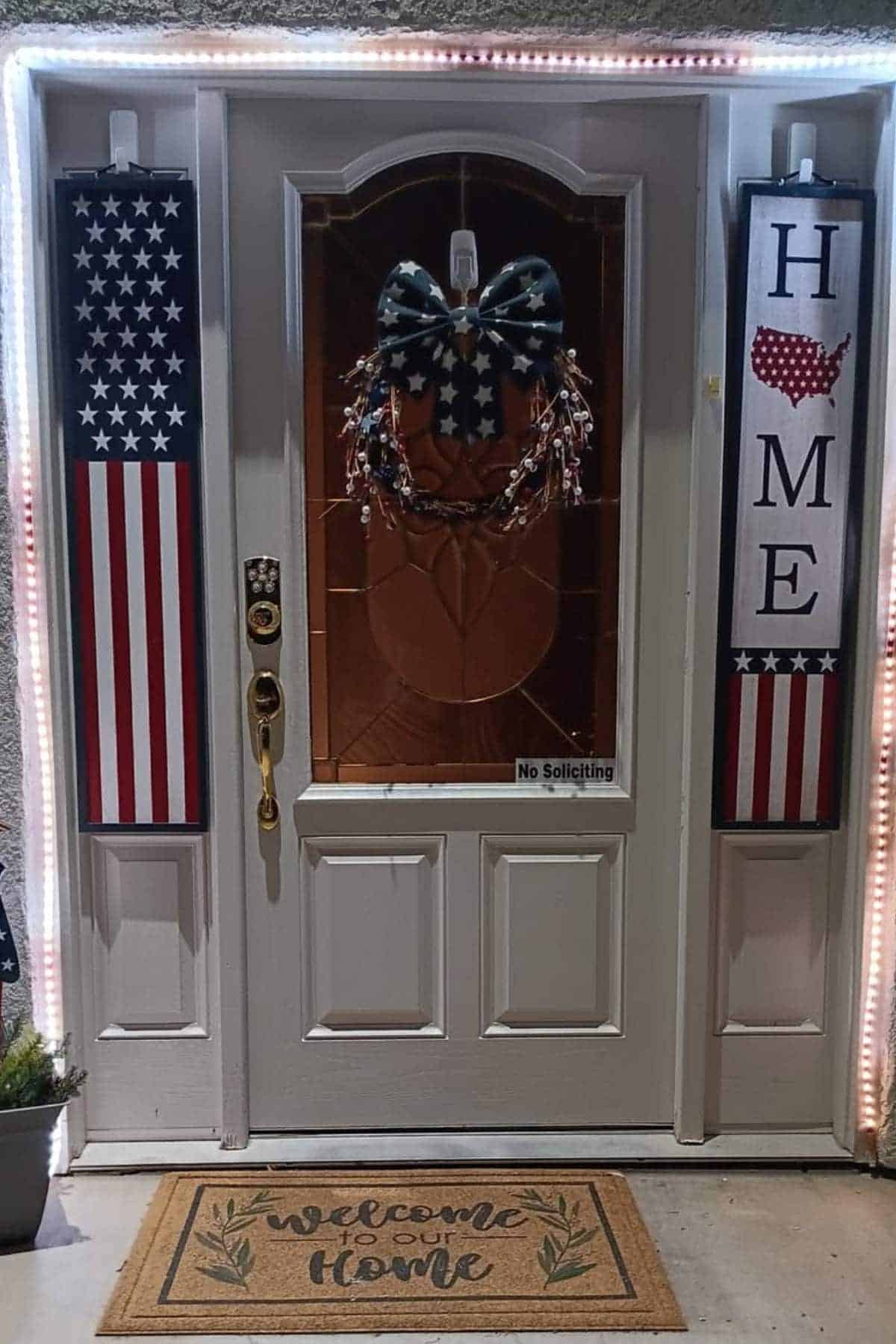 Front door decorated with American flag-themed wreath and banners, one spelling "HOME" with a US map, white trim lights, and a "Welcome to our Home" doormat.