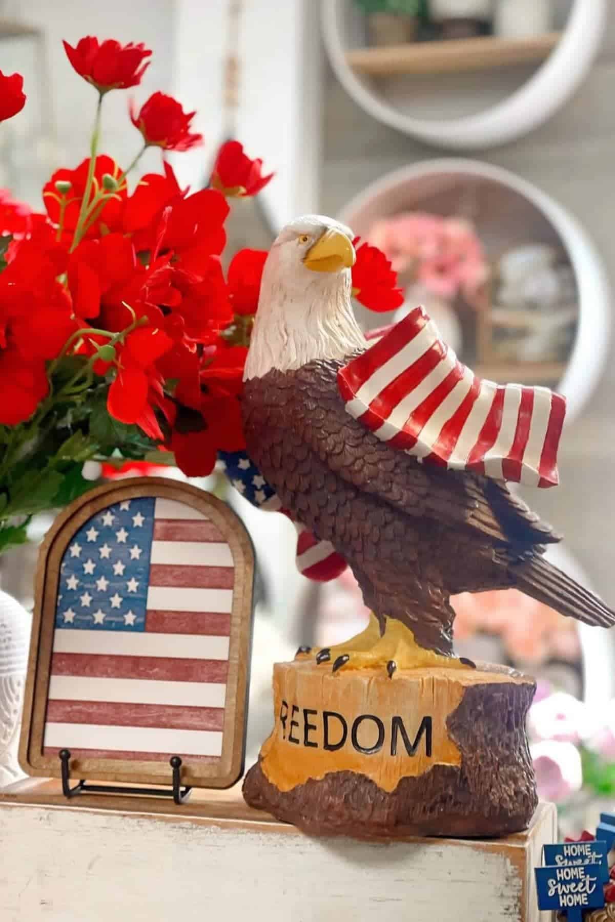 A bald eagle statue draped with an American flag stands on a base labeled "FREEDOM," next to a framed American flag and red flowers.