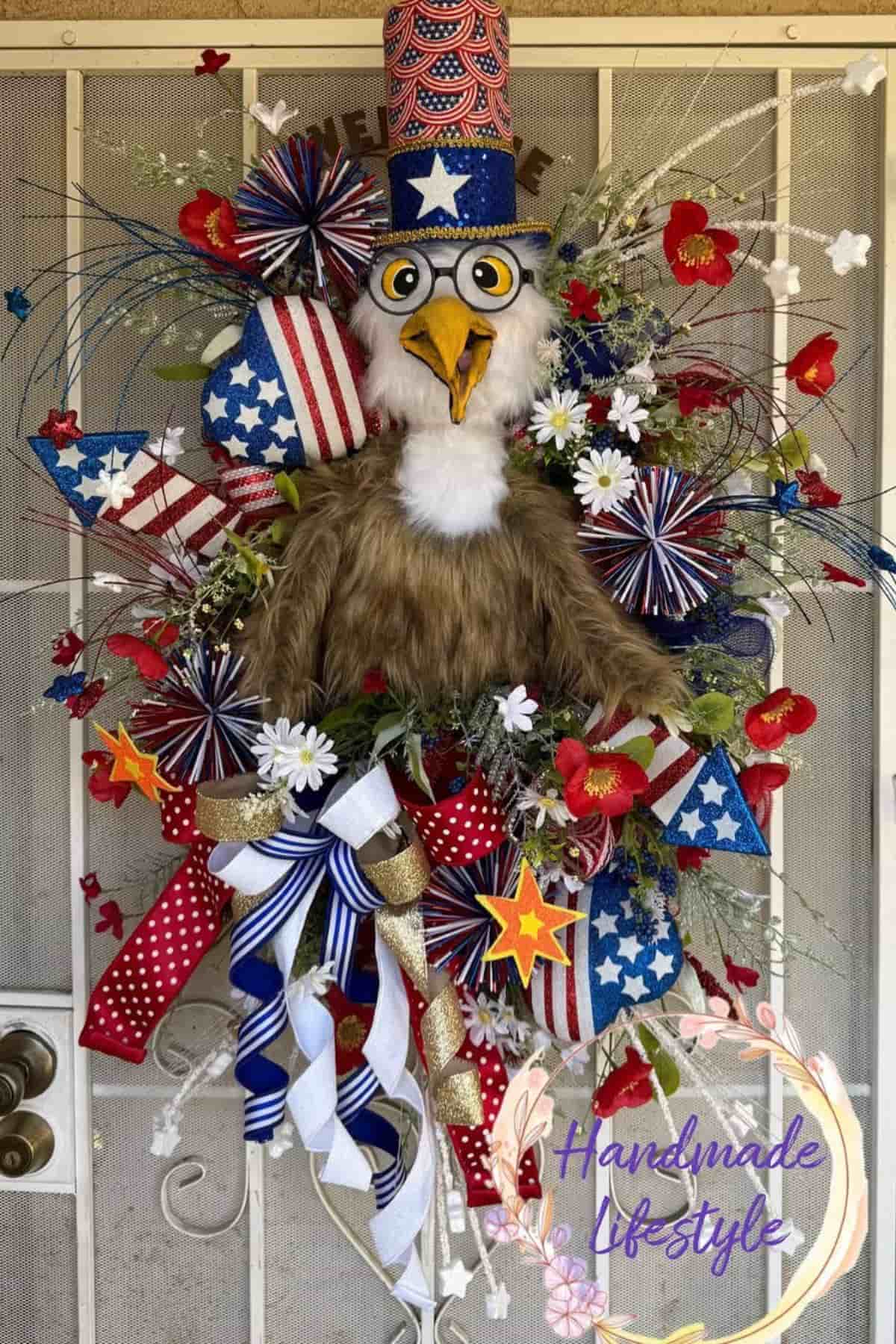 A patriotic wreath featuring a plush eagle wearing a stars-and-stripes hat, surrounded by red, white, and blue ribbons, flowers, and decorative star shapes.
