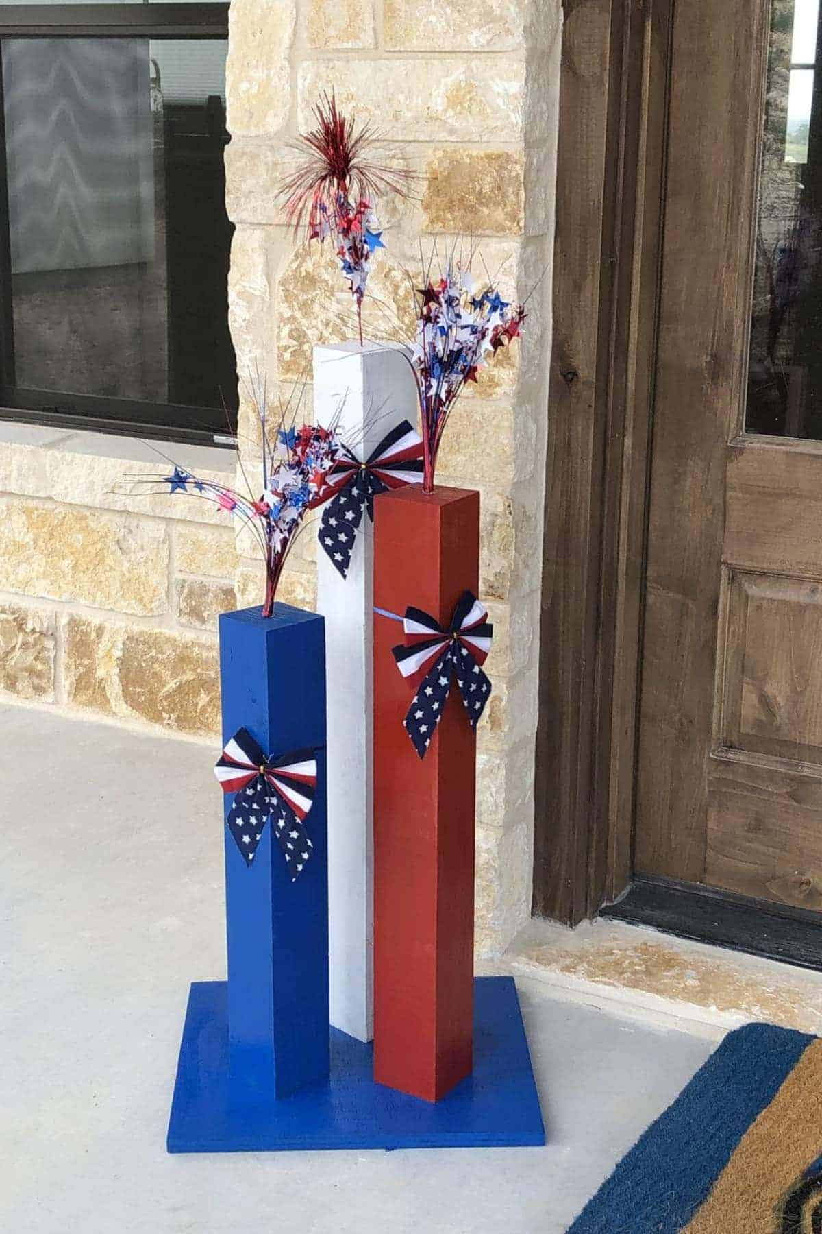 Three tall rectangular wooden blocks painted blue, white, and red stand on a porch, decorated with patriotic bows and star-shaped sprays.