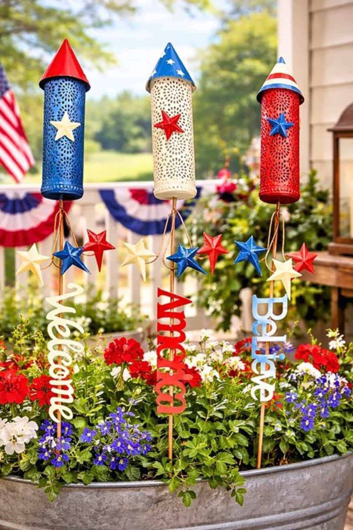 Three rocket-shaped patriotic decorations with "Welcome," "America," and "Believe" stakes in a planter with red, white, and blue flowers.