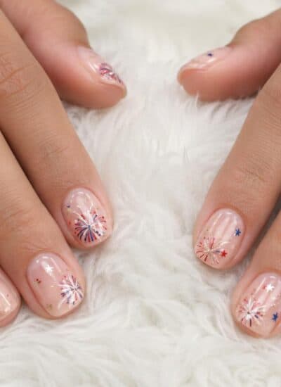 Nails with a sheer nude polish feature red, white, and blue firework designs and gold star accents.