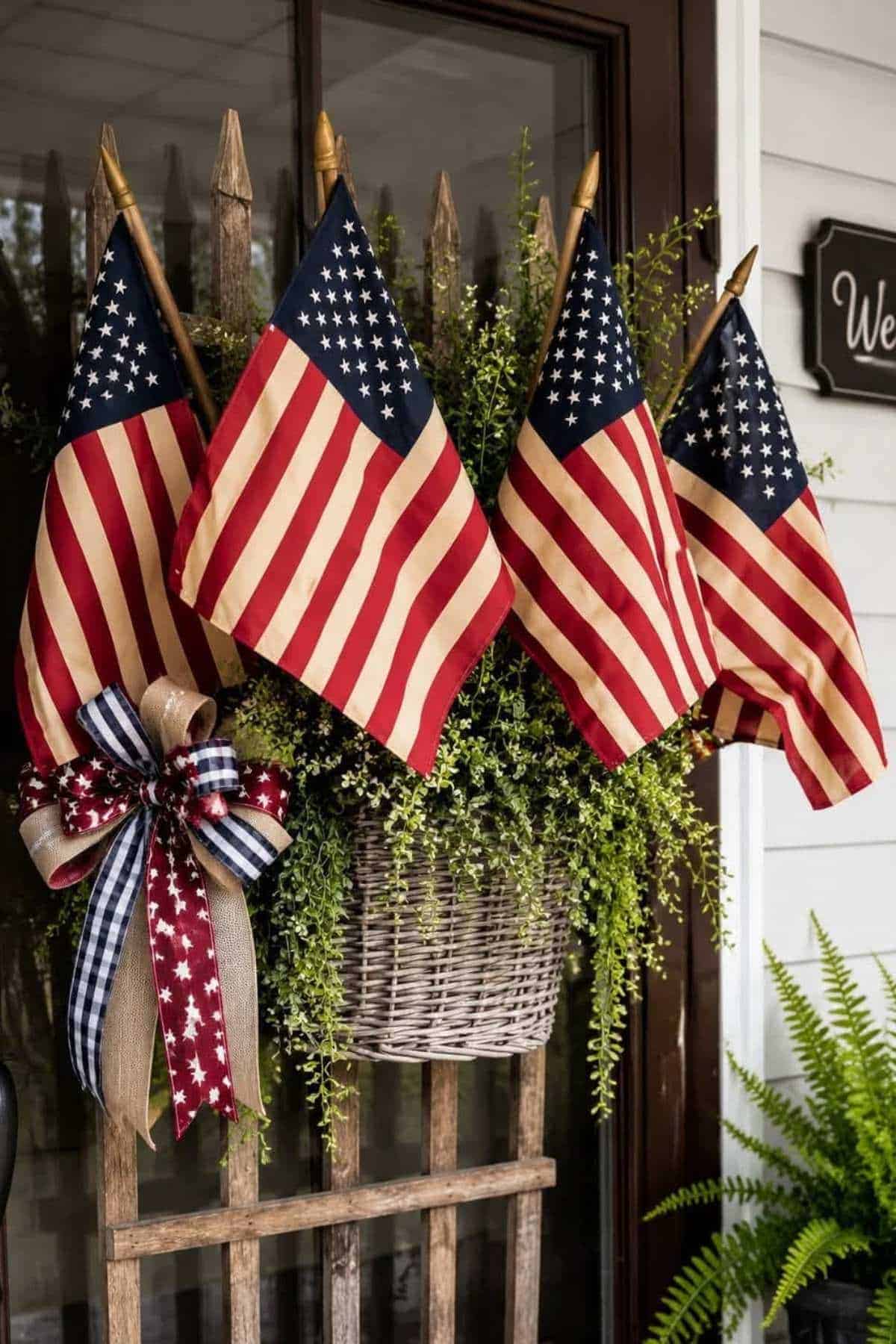 Four American flags hang from a wooden fence in front of a wicker basket overflowing with greenery. A patriotic bow adorns the left side.