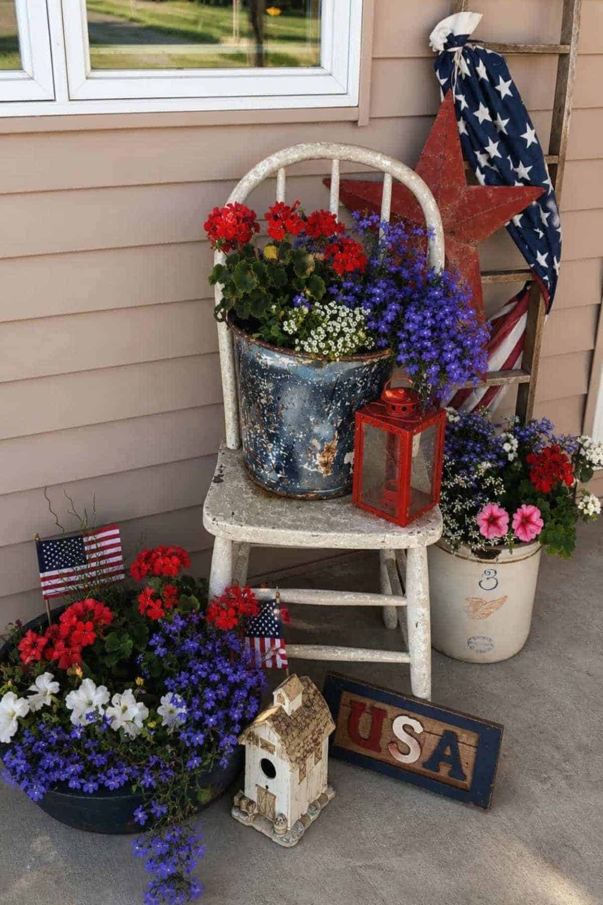 A patriotic porch display with flowers in red, white, and blue, a vintage chair, lantern, and American flag decor.