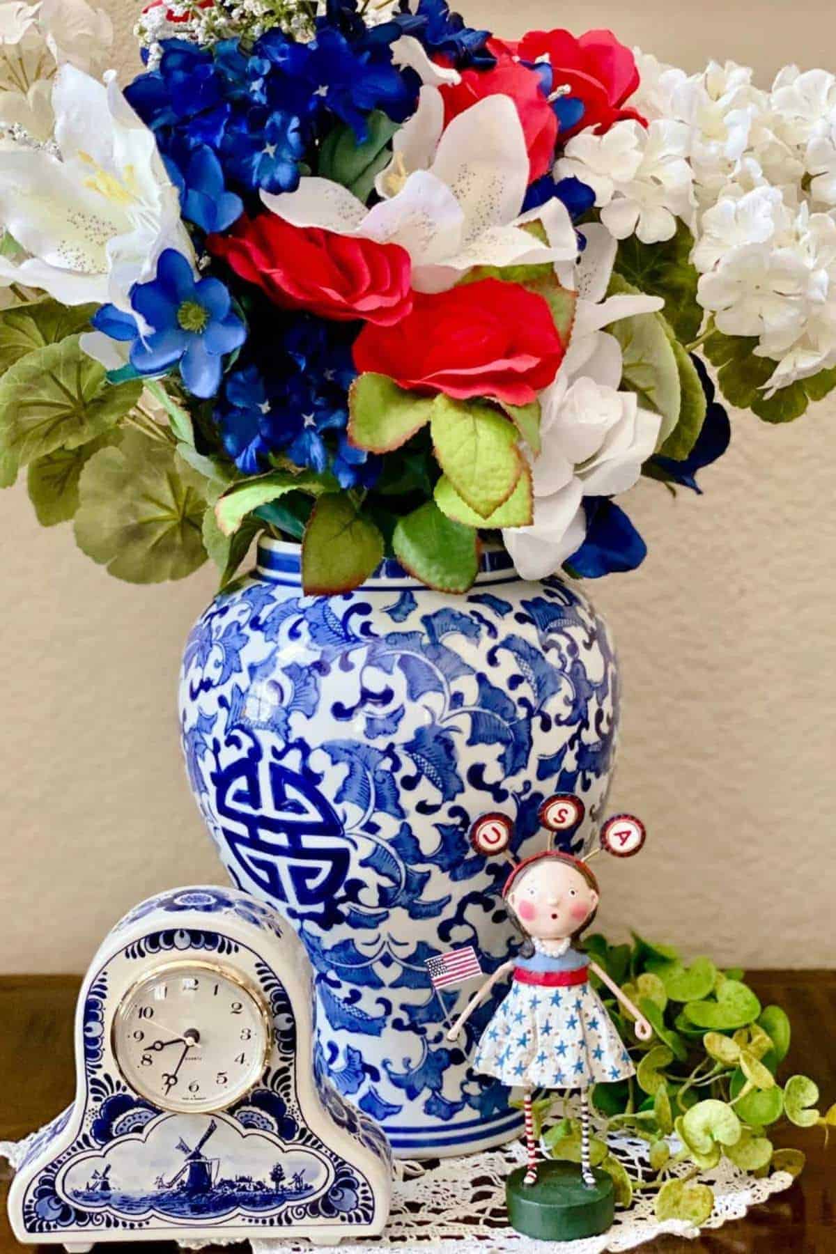 A blue and white vase with red, white, and blue artificial flowers sits beside a decorative clock and a patriotic figurine holding an American flag.
