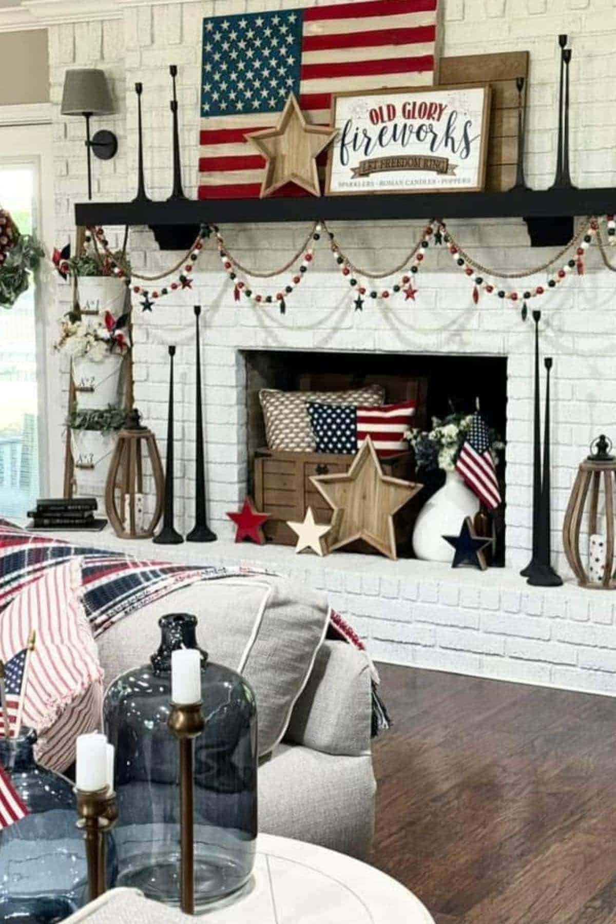 A living room with a white brick fireplace decorated with American flags, wooden stars, patriotic garlands, and red, white, and blue accents for a patriotic theme.