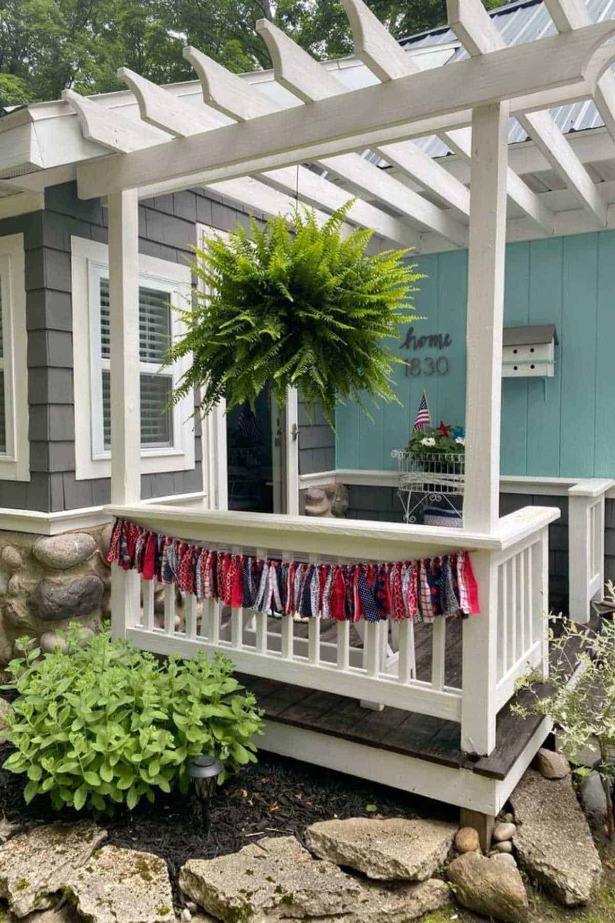 A white pergola covers a porch decorated with a patriotic fabric banner and a hanging fern.