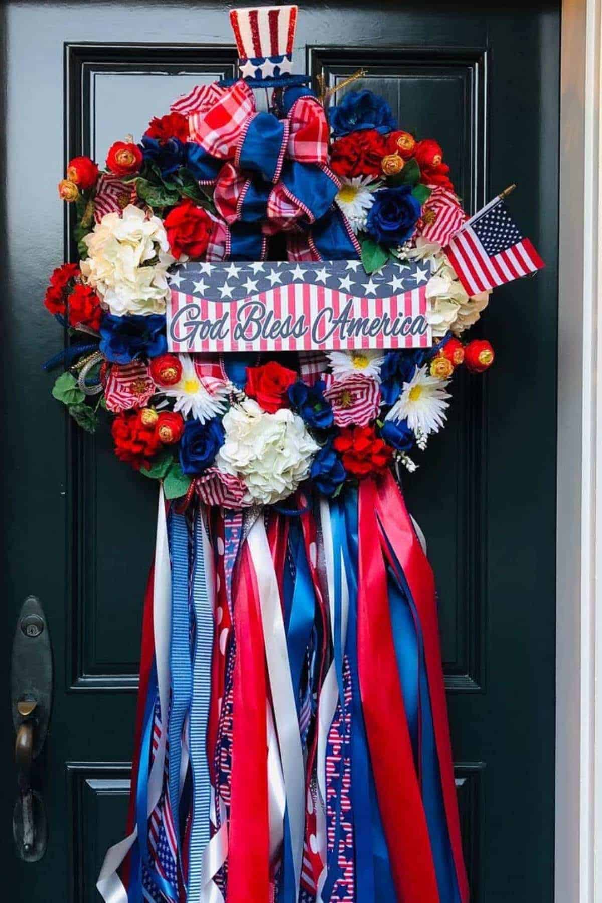A patriotic wreath with red, white, and blue flowers, ribbons, mini American flags, and a "God Bless America" sign hangs on a dark door.