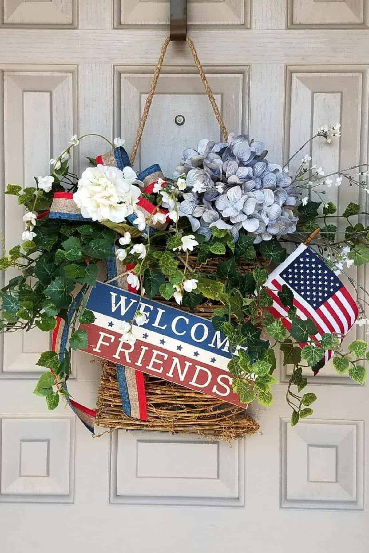 A patriotic wreath with a "WELCOME FRIENDS" sign, flowers, ivy, and an American flag hangs on a white door.