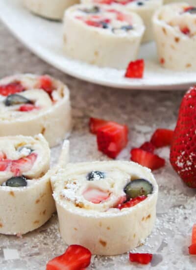 Strawberry and blueberry tortilla roll-ups on a speckled countertop, with sliced strawberries and powdered sugar sprinkled on top.
