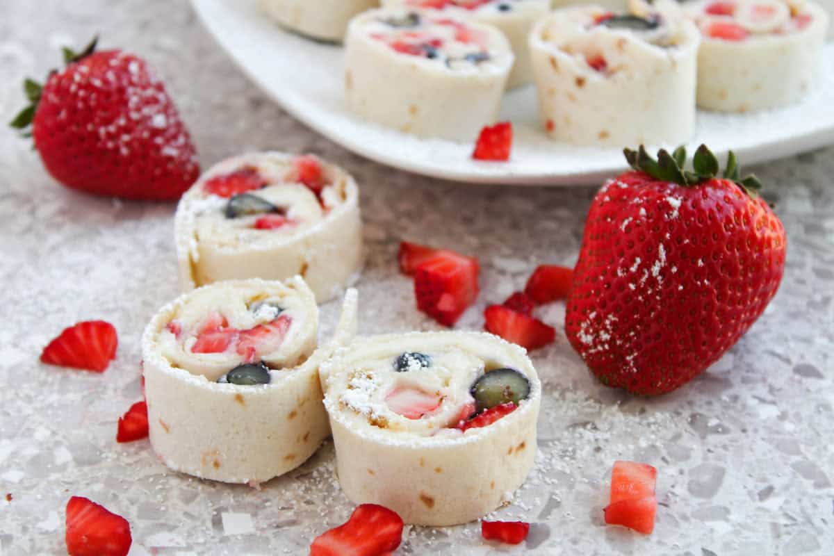 Strawberry and blueberry tortilla roll-ups on a speckled countertop, with sliced strawberries and powdered sugar sprinkled on top.