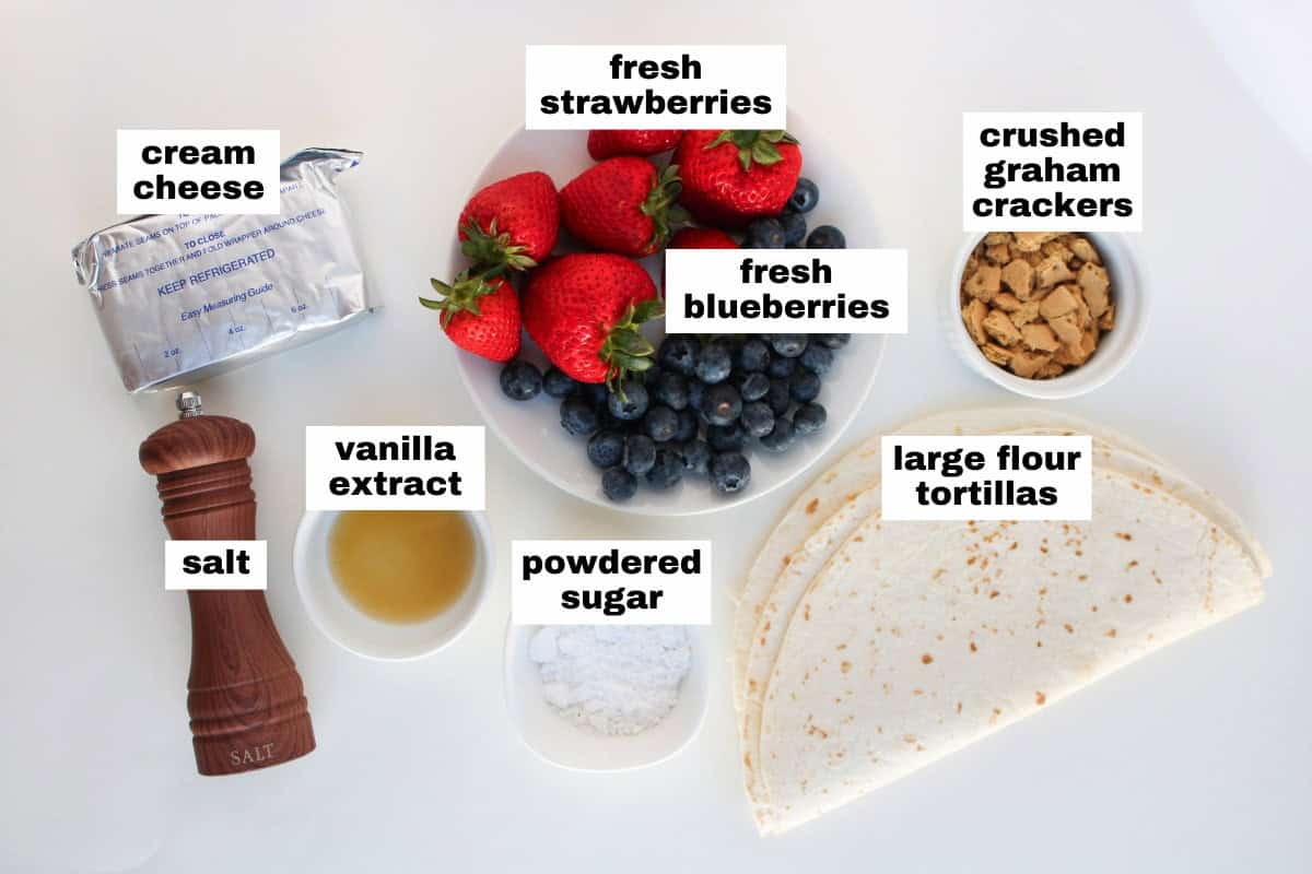 A plate of fresh strawberries and blueberries, cream cheese, crushed graham crackers, large flour tortillas, powdered sugar, vanilla extract, and a salt grinder arranged on a white surface.