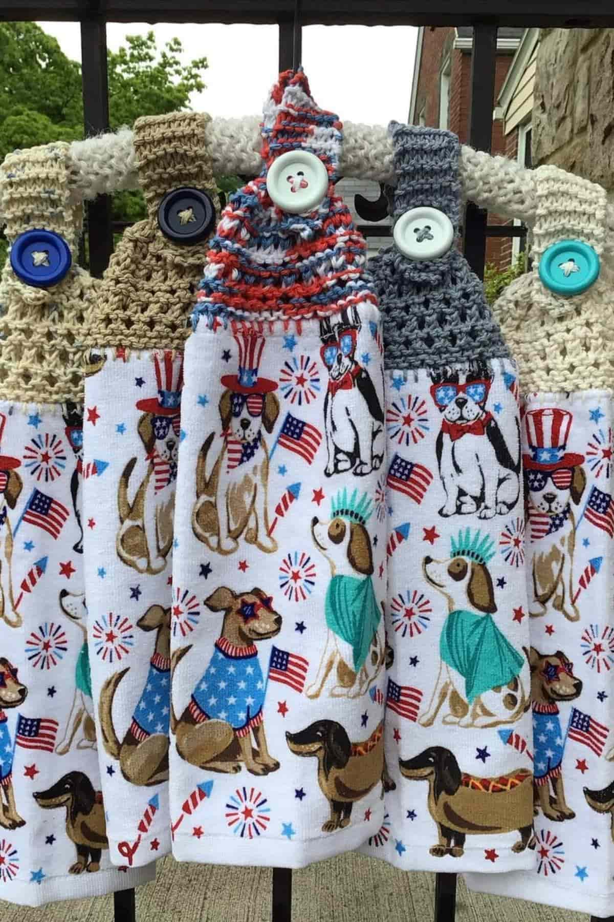Four crocheted hanging towels with buttons, decorated with patriotic-themed dogs, American flags, and Statue of Liberty outfits, displayed on a rack outdoors.