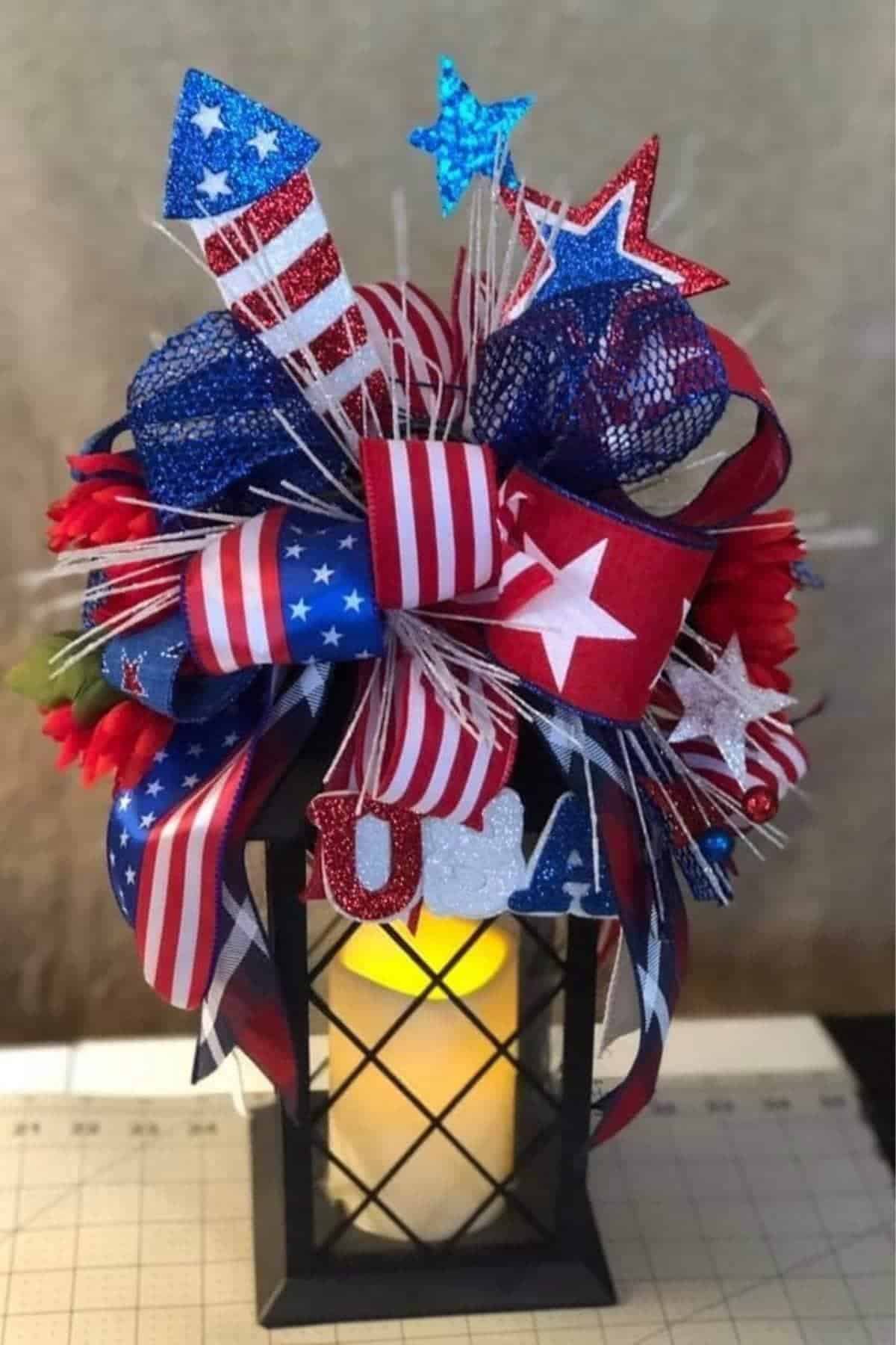 A black lantern with a lit candle is decorated with red, white, and blue patriotic ribbons, glitter stars, and "USA" letters, sitting on a cutting mat.