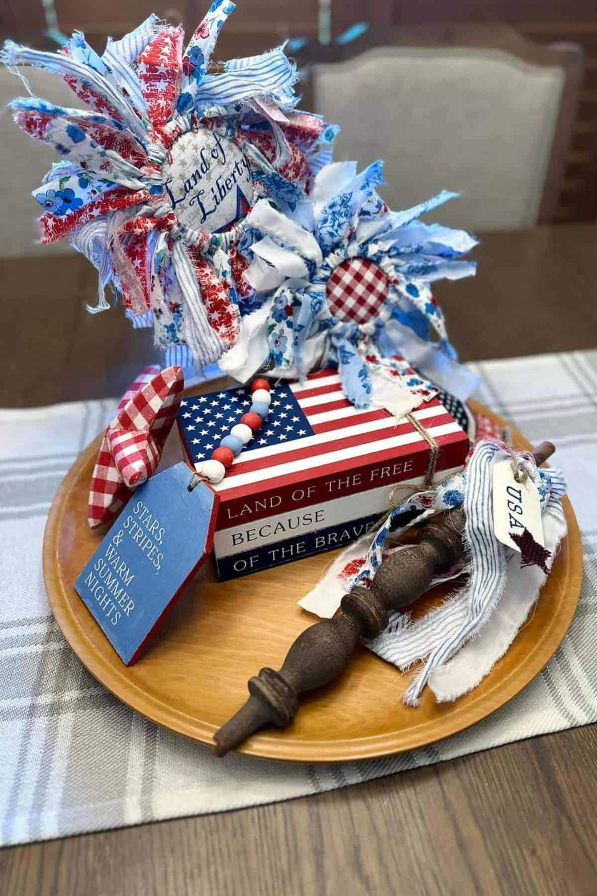 A wooden tray with patriotic-themed decor, including stacked books, fabric bows, painted beads, a wooden spindle, and a USA tag, set on a plaid table runner.