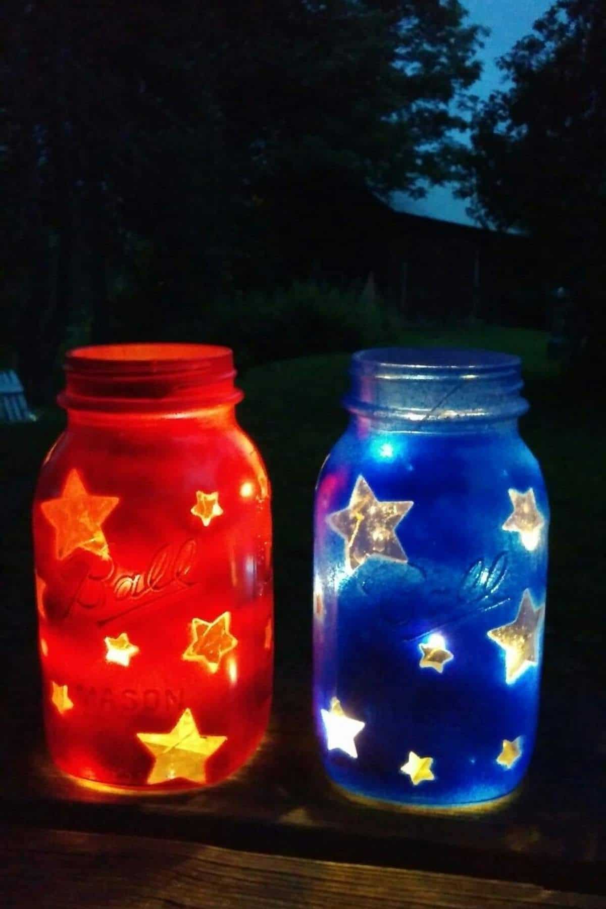 Two mason jars, one red and one blue, are decorated with star cutouts and lit from inside, creating a glowing effect in an outdoor nighttime setting.