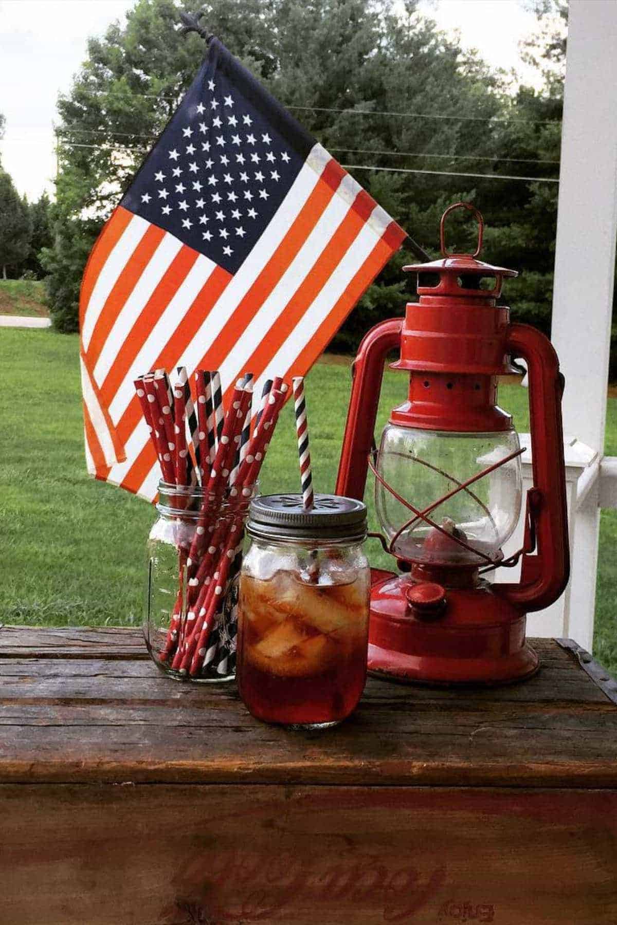 American flag, red lantern, mason jar with drink, and patriotic straws on wooden surface.