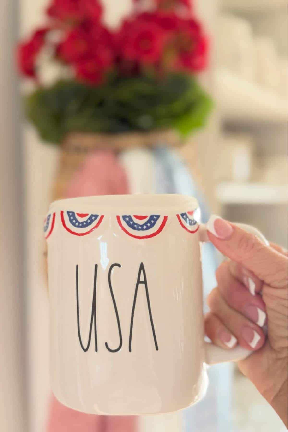 A hand holds a white mug decorated with red, white, and blue bunting and the letters "USA"; blurred red flowers are in the background.