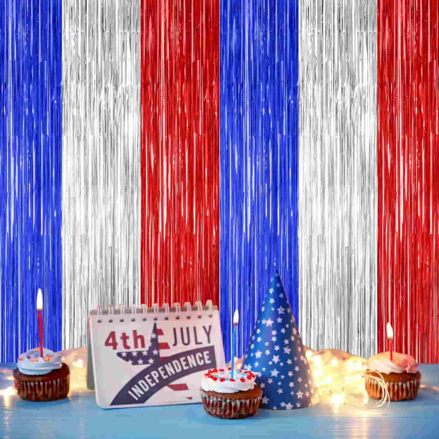 Red, white, and blue tinsel backdrop with cupcakes, lit candles, a 4th of July Independence Day calendar, a blue party hat, and string lights in the foreground.