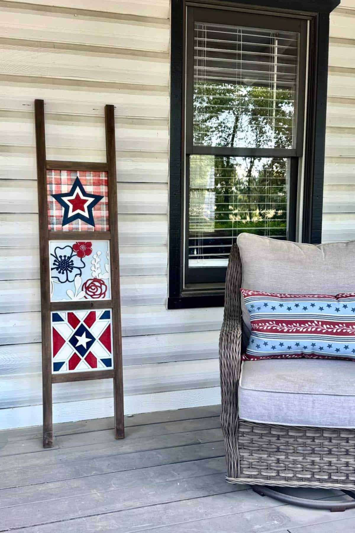 A decorative wooden ladder with patriotic-themed tiles leans against a house wall next to a wicker chair with red, white, and blue pillows on a porch.