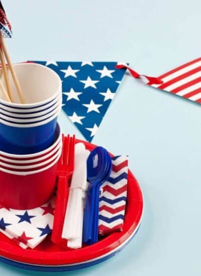 Red, white, and blue party supplies with American flag patterns, including plates, cups, utensils, napkins, and a triangular flag banner, arranged on a light blue background.