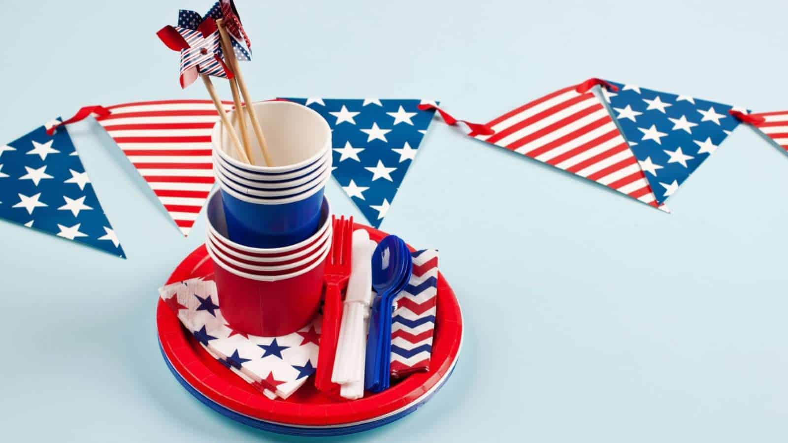 Red, white, and blue party supplies with American flag patterns, including plates, cups, utensils, napkins, and a triangular flag banner, arranged on a light blue background.
