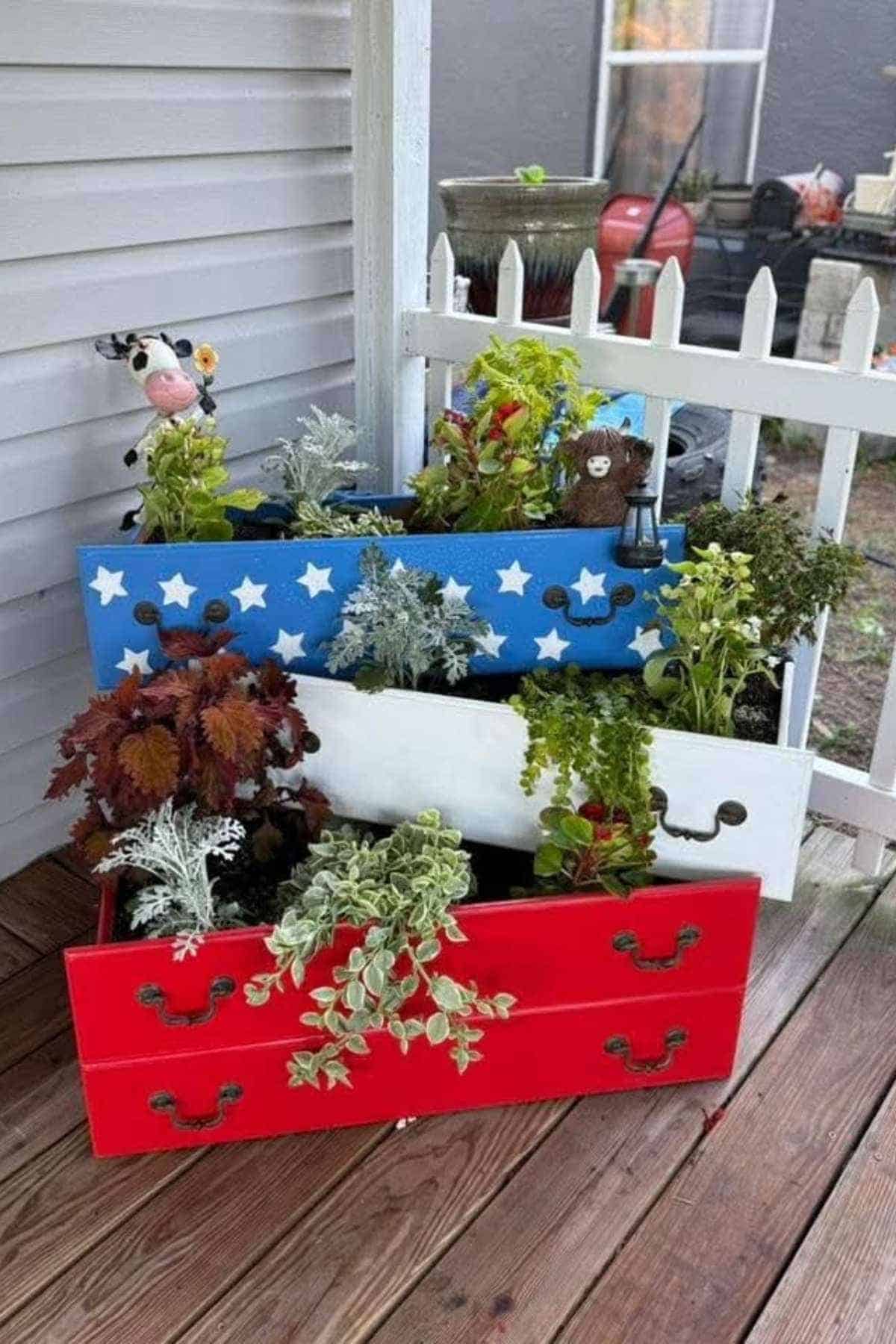 Three painted dresser drawers, red, white, and blue with stars, hold plants on a wooden deck.