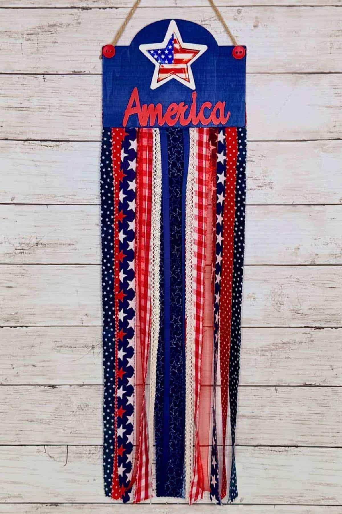 A hanging decoration with "America" text, a star, and long ribbon streamers in red, white, and blue with various patterns, displayed against a white wooden background.