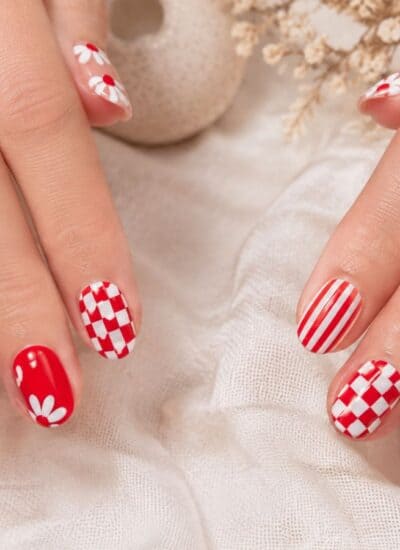 Close-up of hands with manicured nails featuring red and white stripes, checkered patterns, and daisy flower designs on a light fabric background.