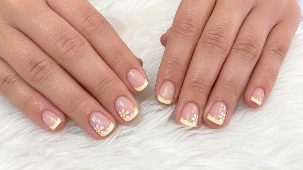 Two hands rest on a white fuzzy surface, displaying short nails with pale yellow French tips and small white daisy designs on some nails.