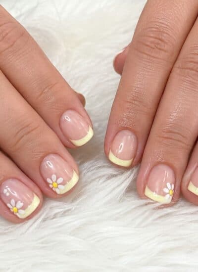 Two hands rest on a white fuzzy surface, displaying short nails with pale yellow French tips and small white daisy designs on some nails.