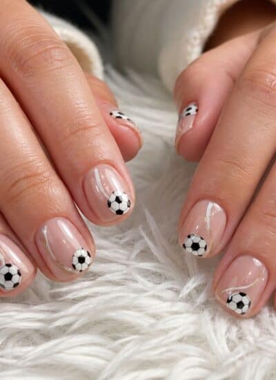 Close-up of hands with manicured nails featuring small black and white soccer balls as nail art, resting on a white furry surface.