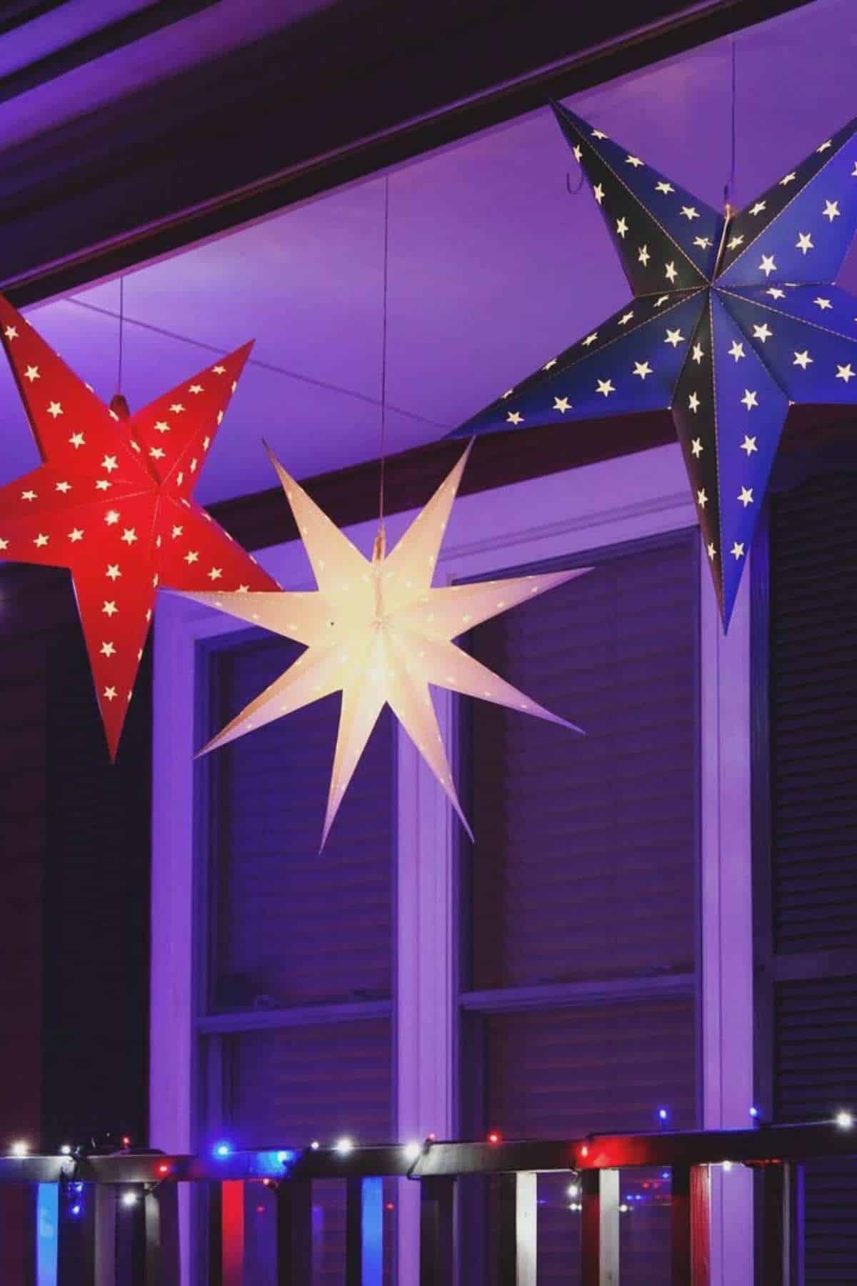 Three large paper star lanterns, red, white, and blue with white stars, hang under a purple-lit porch ceiling above a window.