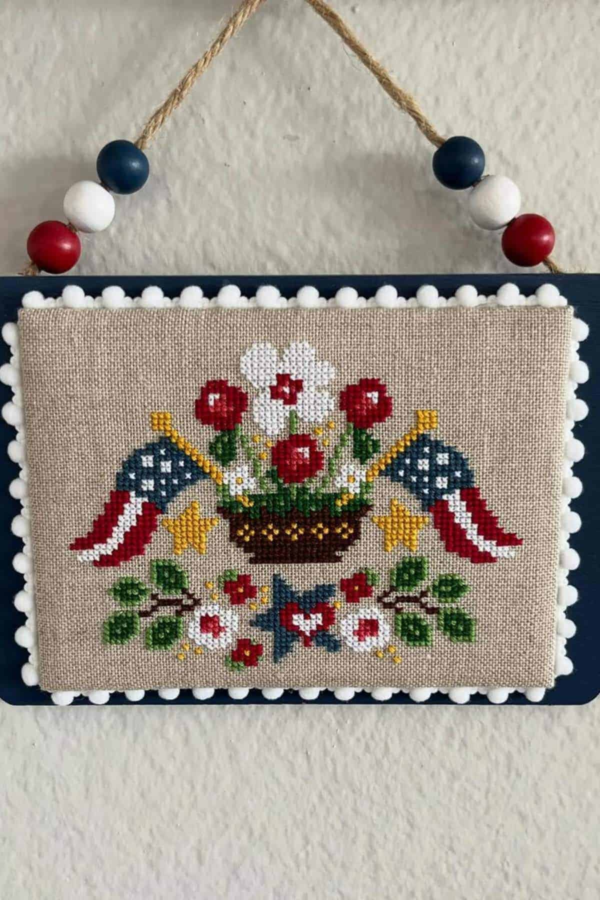A cross-stitch design features a basket of flowers, two American flags, and star accents, mounted on a fabric board with pom-pom trim and a twine hanger decorated with red, white, and blue beads.