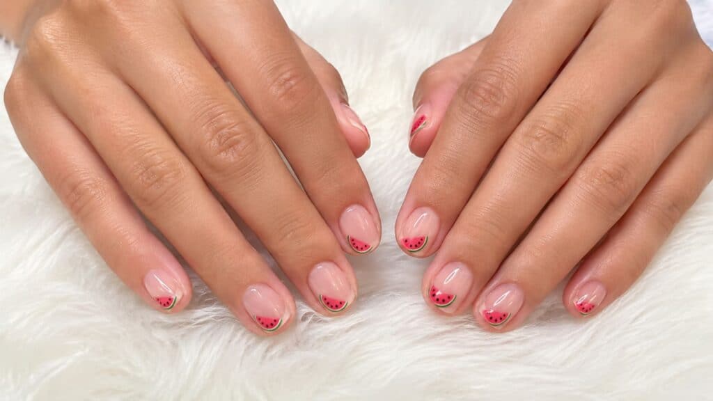 Two hands with a light pink manicure featuring small watermelon slice designs on the tips, resting on a white furry surface.