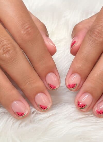 Two hands with a light pink manicure featuring small watermelon slice designs on the tips, resting on a white furry surface.