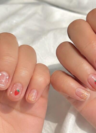 Two hands with short, natural nails featuring playful nail art, including strawberries, clouds, stars, and sun rays on a light pink base.