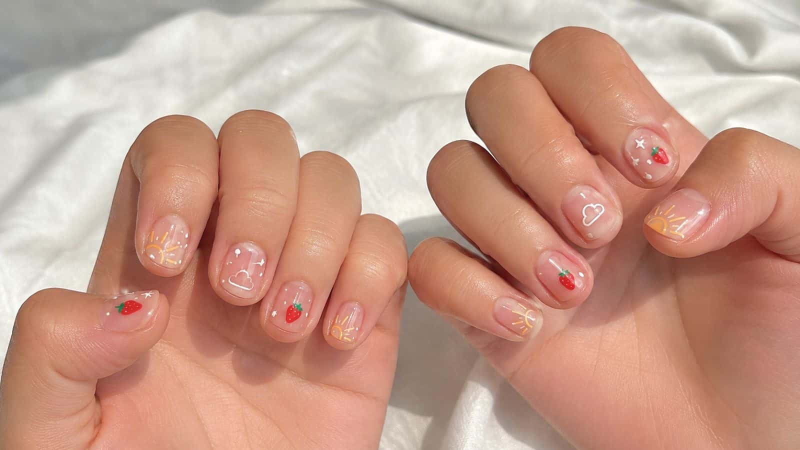 Two hands with short, natural nails featuring playful nail art, including strawberries, clouds, stars, and sun rays on a light pink base.