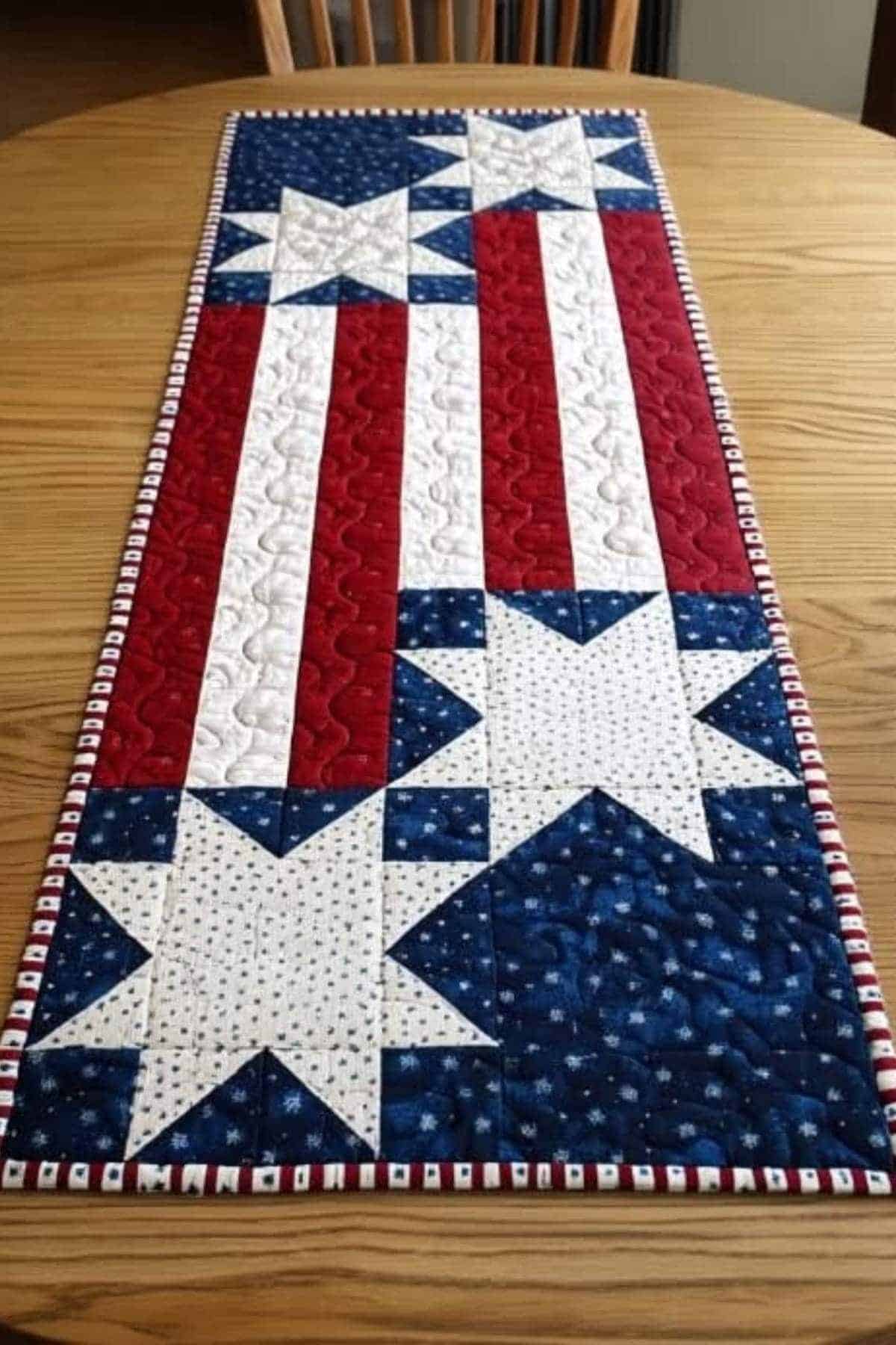 A quilted table runner with red, white, and blue stripes and white stars, resembling an American flag, displayed on a wooden table.